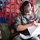 Saving Lives in the Battlespace A female military personnel member wearing a flight suit and aviation headset is seated inside what appears to be a military transport aircraft, reviewing documents. Her name tag reads 'KIM CARR' and her flight suit displays patches indicating affiliation with the Air National Guard. A military-issue bag labeled 'CARROLL' is visible beside her.