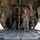Saving Lives in the Battlespace The image shows the interior of a large military cargo aircraft, viewed from the open rear ramp looking inward. Two personnel in olive drab flight suits are visible inside the cargo bay — one standing and looking upward while wearing a headset, and one kneeling on the floor. The aircraft's structural framework, cargo hooks, tie-down rings, and mechanical systems are clearly visible along the ceiling and walls.