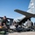 Saving Lives in the Battlespace Military personnel are conducting an aeromedical evacuation exercise or operation on a tarmac beside a Texas Air National Guard C-130 Hercules aircraft (tail number 31456). Multiple simulated casualties on litters/stretchers are being attended to by flight crew and medical personnel. The scene depicts a mass casualty loading drill or real-world medical evacuation scenario.
