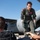 Saving Lives in the Battlespace A female military aviator in an olive green flight suit is leaning forward and communicating with personnel on the ground near a military transport aircraft. The aircraft in the background has partial text visible reading 'S AIR GUARD', indicating a National Guard unit. The scene appears to be on an active flight line or tarmac during a training or operational exercise.