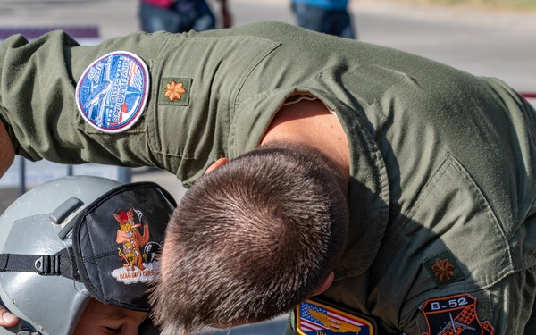 A military aviator in an olive green flight suit is fitting a pilot's helmet onto a young child at what appears to be an air show or military open day. The airman's patches indicate affiliation with the B-52 program and Air Force Reserve, with a name tape reading 'TIM HUMPAL' and a Czech flag patch visible. The child is smiling, suggesting this is an interactive public engagement activity.