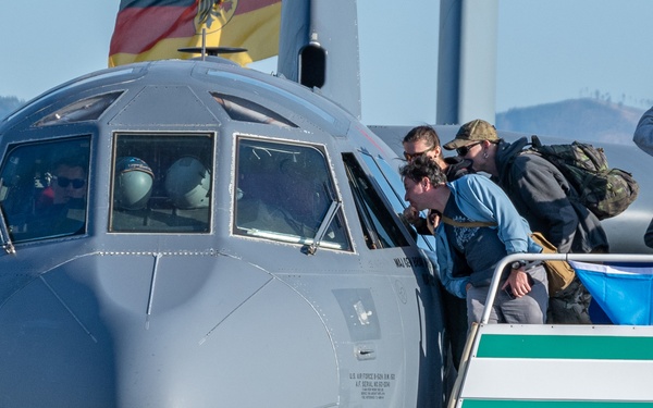 A U.S. Air Force military transport aircraft, identifiable as a C-130 Hercules, is shown on the ground at what appears to be an air show. Civilians and visitors are gathered on a boarding stairway/platform alongside the aircraft, with some leaning over to look into the cockpit. A German flag is visible in the background, suggesting this event is taking place in Germany.