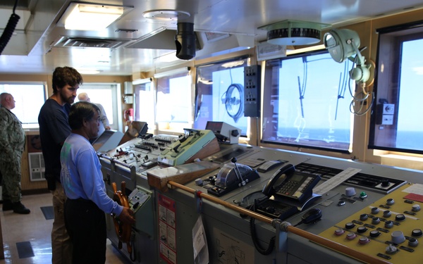 The image shows the bridge or control room of a naval or research vessel, with multiple personnel operating navigation and control equipment. Several large monitors display external camera feeds showing the ship's deck and equipment. The space is densely packed with control panels, switches, and communication devices.