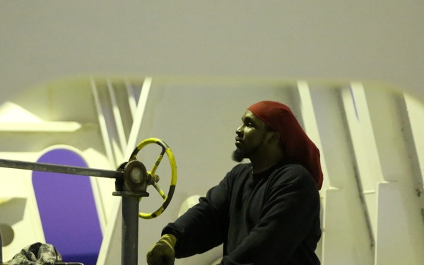 The image is captured through a rounded rectangular porthole or access hatch, revealing a worker operating machinery in what appears to be an industrial or maritime environment. The man, wearing a red beanie and dark hoodie with yellow work gloves, is handling mechanical equipment including a yellow handwheel valve. The background shows white-painted walls with purple geometric markings, suggesting a ship or large industrial facility.