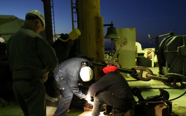 Three workers are performing maintenance or repair work on what appears to be the deck of a ship or offshore platform at night. Two workers are crouched down working on equipment near a heavy chain and mooring hardware, while a third worker in a hard hat stands observing. A bright work light illuminates the immediate work area against the dark night sky.