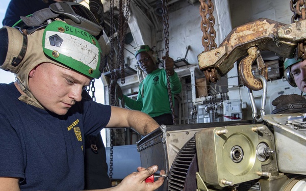 Military personnel, likely U.S. Navy sailors, are performing maintenance or repair work on a piece of heavy mechanical equipment in what appears to be a ship's hangar bay or maintenance deck. The primary worker in the foreground is kneeling and using a hand tool on the machinery while wearing a cranial (flight deck helmet) with green trim and hearing protection. Additional crew members observe or assist in the background.
