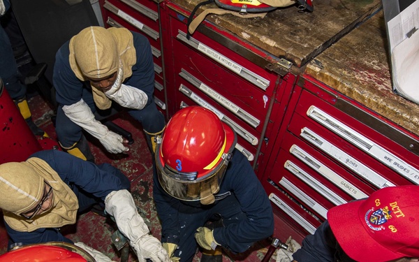 A group of naval personnel, likely U.S. Navy sailors, are engaged in a damage control training exercise aboard a ship. They are working on a pipe repair task, using plugging and patching techniques on what appears to be a simulated pipe leak. Personnel are wearing protective gear including red helmets, flash hoods, and work gloves.