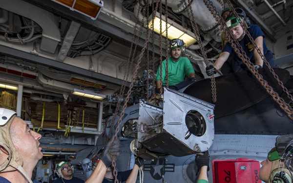 Military personnel, likely U.S. Navy sailors, are performing maintenance or component removal on a helicopter inside a ship's hangar bay. Several crew members wearing green shirts, helmets, and hearing protection are working together using chains and hoisting equipment to lift or remove a large component from the aircraft. A red box labeled '706' is visible on the side of the helicopter.
