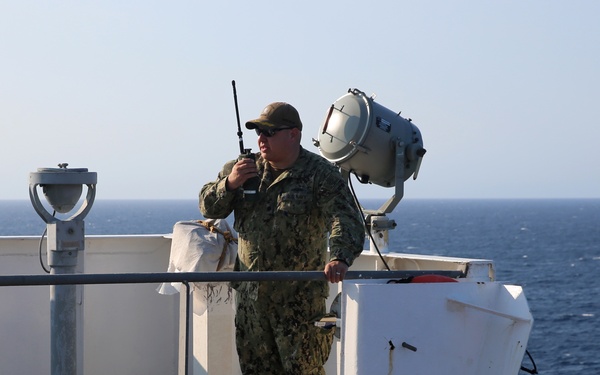 A military personnel member in camouflage uniform stands on the elevated bridge wing or observation deck of a naval or military vessel at sea. The individual is holding a handheld radio/walkie-talkie to their mouth, apparently communicating while on watch duty. The vessel is underway on open blue ocean under clear skies.