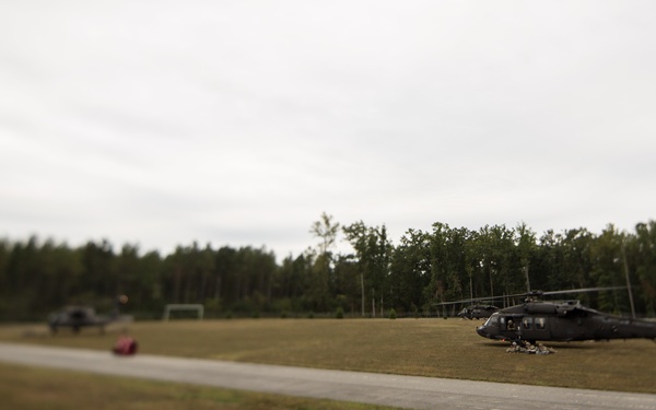 The image shows at least two dark military helicopters, consistent with UH-60 Black Hawks, parked on or near a paved strip in an open grassy field. Personnel and equipment appear to be gathered near the closer helicopter on the right side. The scene appears to be a field landing zone or training area bordered by a tree line.