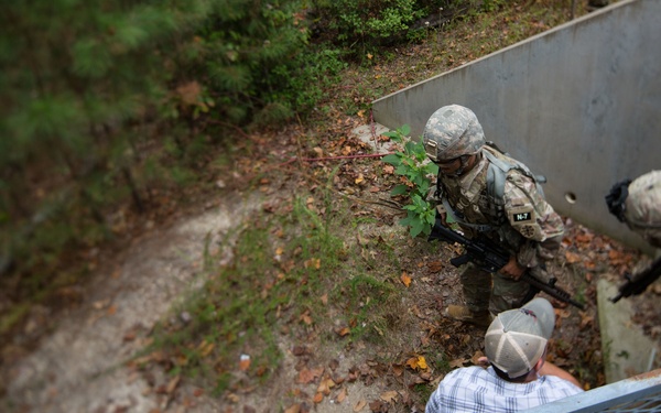 An overhead view shows two armed soldiers in military camouflage uniforms and helmets interacting with a civilian near a concrete structure or stairwell entrance. The soldiers appear to be conducting a training exercise or security operation, with one soldier visibly labeled 'N-7' on their gear. The scene is set in a wooded outdoor environment with fallen leaves on the ground.
