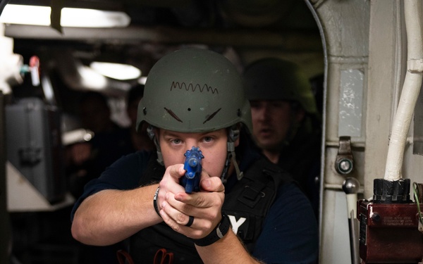 A military or naval personnel member is conducting a training exercise aboard what appears to be a naval vessel. The individual is wearing a helmet, body armor, and holding a blue training/simulation pistol in a two-handed grip while moving through a watertight door/hatch. A second personnel member is visible behind them in the narrow corridor.