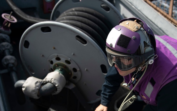 A naval personnel member wearing a purple vest and helmet with a face shield is working on fuel or fluid transfer equipment aboard what appears to be a military vessel. The individual is manipulating a purple-colored valve or fitting connected to a hose system. The purple color coding of both the worker's gear and the equipment is consistent with aviation fuel (JP-5) handling operations on a naval vessel.