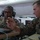 Two U.S. Marines in digital camouflage uniforms are working together at a computer workstation inside what appears to be a military command tent. One Marine wearing glasses and a headset is seated at the workstation, while the other Marine, whose name tape reads 'DRONE' and bears a staff sergeant chevron, points toward the screen. The setting suggests a tactical operations or command and control environment.