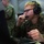 A young military personnel wearing MARPAT camouflage uniform, glasses, and a communications headset is focused on a laptop computer in what appears to be a command operations tent. At least two other uniformed personnel are visible in the background. The setting suggests a tactical operations center or field command post environment.