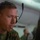 A male military personnel wearing digital camouflage (MARPAT) uniform is focused on a monitor screen in what appears to be a command or operations center. He is equipped with a communications headset featuring a boom microphone, and has protective eyewear pushed up on his forehead. The image is a close-up portrait-style shot with a blurred background showing at least one other person.