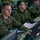 Multiple military personnel in MARPAT (Marine pattern) camouflage uniforms are working at computer workstations in what appears to be a command operations center or tactical operations center (TOC). The foreground subject is focused on a Dell laptop while a large monitor is visible to the right. Several other service members are visible working at similar stations in the background.