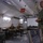 Military personnel in camouflage uniforms are gathered inside a large field tent for what appears to be a briefing or planning session. A standing individual addresses three seated personnel, with a projection screen visible in the background. A red sign reading 'Future Operations' is suspended from the tent ceiling, indicating this is a military command or operations planning environment.