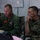 Two U.S. Marine Corps personnel are seated at a workstation in what appears to be a command operations center or tactical operations center (TOC). The individual on the left is wearing a green flight suit with USMC patches and aviator wings, while the individual on the right is in MARPAT digital camouflage utilities with lieutenant colonel oak leaf insignia. Both appear to be reviewing information on a screen during what appears to be a briefing or operational planning session.