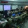 Military personnel, identifiable as U.S. Marines by their digital camouflage uniforms and visible Eagle Globe and Anchor emblem on a laptop screen, are operating inside a large command tent. The space is configured as a tactical operations center (TOC) with multiple workstations, laptops, desktop monitors, and large projection screens. Personnel appear to be engaged in a command and control exercise or real-time operational coordination.