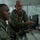 Two military personnel in digital camouflage uniforms and tactical vests are working at a computer workstation inside a tent or temporary command structure. One individual is seated at the keyboard while the other stands behind and points at the monitor screen, appearing to provide instruction or guidance. The setting suggests a field command post or operations center environment.