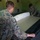 Two individuals in U.S. Marine Corps woodland MARPAT camouflage uniforms are working together to set up or assemble a cot or field bed inside a building with green walls. They appear to be placing a mattress or pad onto a folding cot frame. The setting appears to be a military facility or barracks based on the visible 'Emergency Exit' signage on the wall.