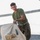 A young male service member wearing USMC MARPAT woodland camouflage trousers and an olive drab green t-shirt is carrying a large tan/khaki military-style duffel or equipment bag on a concrete surface. He appears to be in the process of moving or transporting the bag, looking toward the camera. The setting appears to be an outdoor paved area in bright daylight conditions.