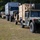 A military convoy of vehicles is lined up on a grassy field, likely at a training base or staging area surrounded by pine trees. The convoy includes multiple HMMWVs (Humvees), a large military cargo truck hauling a shipping container marked 'AC2S', and a vehicle carrying a cable reel spool. Personnel are visible inside and around the vehicles, suggesting preparation for movement or a field exercise.