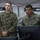 Two U.S. Marines in MARPAT camouflage uniforms are standing in what appears to be a command operations center or tactical operations center (TOC). Both wear name tapes and rank insignia, with a whiteboard labeled 'RESTORATION' visible in the background. They appear to be reviewing information on computer monitors.