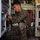 A U.S. Marine in MARPAT digital camouflage uniform is performing maintenance or technical work inside what appears to be a military electronic equipment shelter or vehicle interior. He is crouched between two equipment racks filled with electronic components, cables, and hardware. The image captures focused, hands-on technical work in a confined military equipment space.