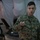 A U.S. Marine identified by name tape as 'Valencia' is conducting a briefing or presentation inside what appears to be a military command tent. He is holding a laser pointer or clicker and appears to be speaking. Additional personnel are visible in the background, one wearing a headset.