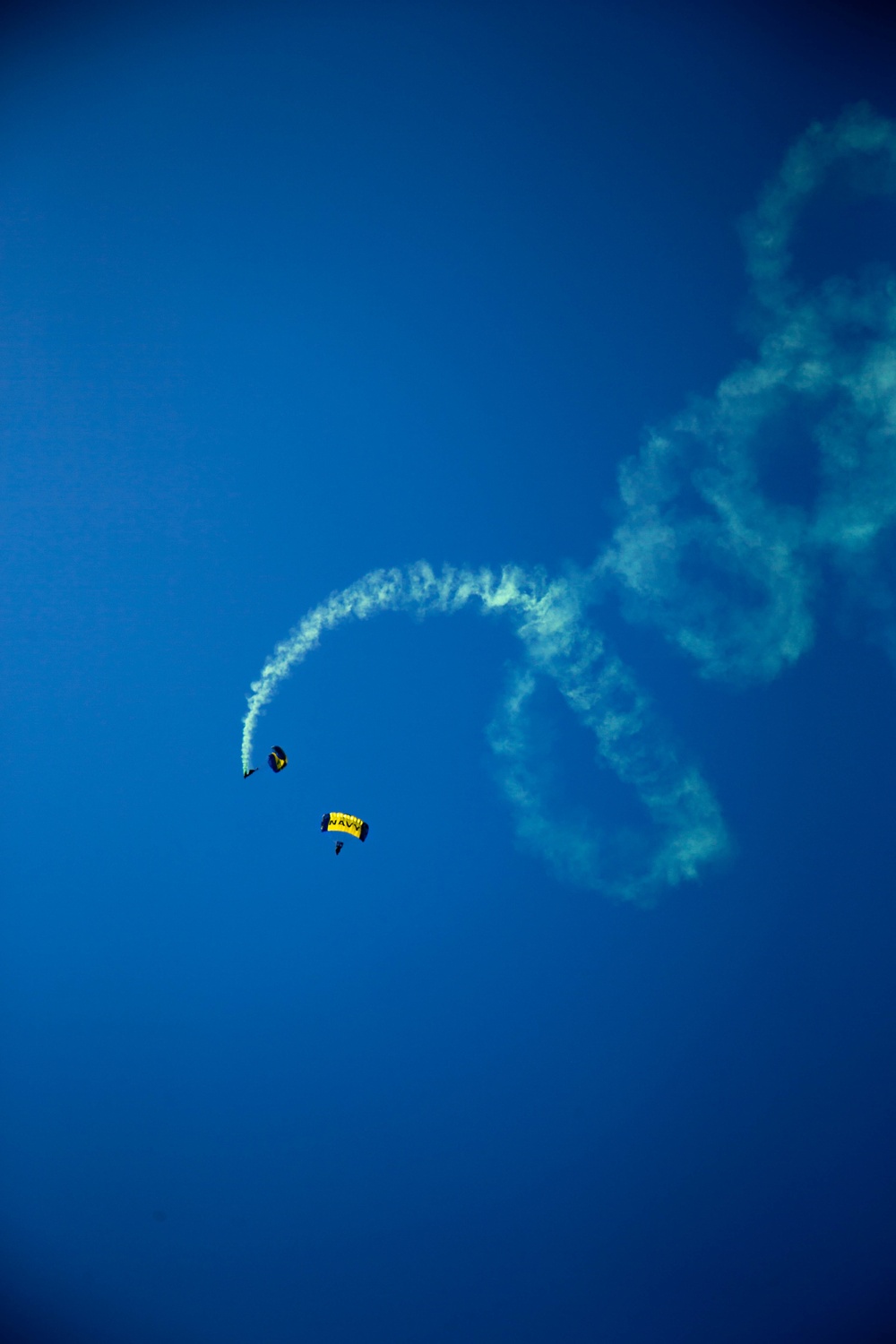 Two skydivers are descending against a clear deep blue sky, with one trailing white smoke in a curved arc pattern. The lower parachute is yellow and bears the text 'NAVY', indicating this is a U.S. Navy parachute demonstration team performance. The smoke trail forms a sweeping circular pattern, suggesting a coordinated aerial display maneuver.