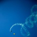 Two skydivers are descending against a clear deep blue sky, with one trailing white smoke in a curved arc pattern. The lower parachute is yellow and bears the text 'NAVY', indicating this is a U.S. Navy parachute demonstration team performance. The smoke trail forms a sweeping circular pattern, suggesting a coordinated aerial display maneuver.