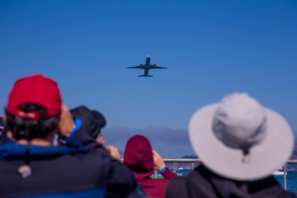 A group of spectators watches a commercial aircraft flying overhead against a clear blue sky. The scene appears to be near a waterfront or bay area, with a bridge and water visible in the background. The viewers are photographed from behind, creating a perspective that draws the eye upward toward the plane.