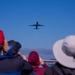 A group of spectators watches a commercial aircraft flying overhead against a clear blue sky. The scene appears to be near a waterfront or bay area, with a bridge and water visible in the background. The viewers are photographed from behind, creating a perspective that draws the eye upward toward the plane.