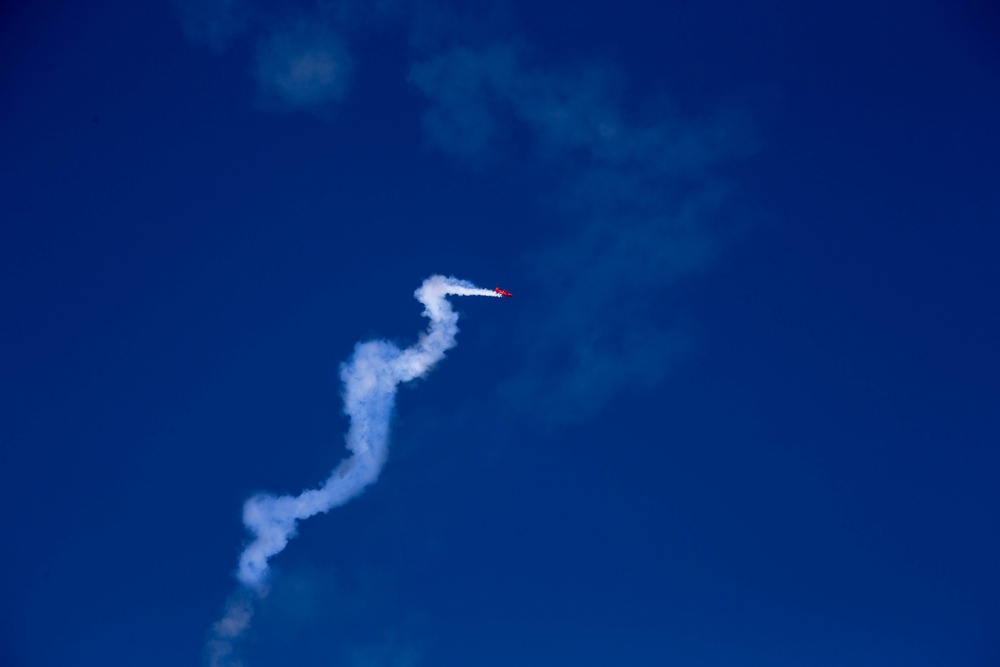 A small red aircraft is performing aerobatic maneuvers against a deep blue sky, leaving a winding white smoke trail behind it. The serpentine smoke trail indicates the aircraft has executed multiple direction changes, consistent with an airshow or aerobatic display. The image is taken from a ground-level perspective looking upward.