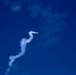 A small red aircraft is performing aerobatic maneuvers against a deep blue sky, leaving a winding white smoke trail behind it. The serpentine smoke trail indicates the aircraft has executed multiple direction changes, consistent with an airshow or aerobatic display. The image is taken from a ground-level perspective looking upward.