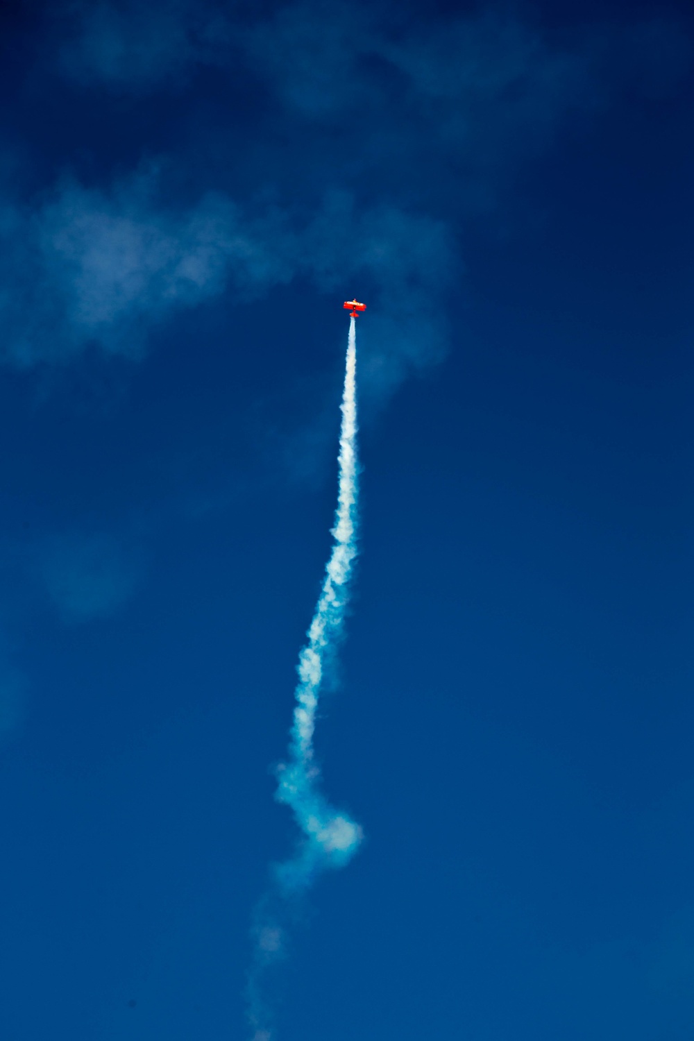 A small red aircraft is performing an aerobatic maneuver, climbing nearly vertically against a deep blue sky. The plane is emitting a white smoke trail that curves slightly, indicating a recent directional change during the performance. This appears to be an airshow or aerobatic display.