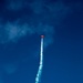 A small red aircraft is performing an aerobatic maneuver, climbing nearly vertically against a deep blue sky. The plane is emitting a white smoke trail that curves slightly, indicating a recent directional change during the performance. This appears to be an airshow or aerobatic display.