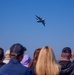 A crowd of spectators watches a large military transport aircraft performing a banking maneuver at an airshow against a clear blue sky. The aircraft appears to be a C-17 Globemaster III or similar heavy military transport, captured mid-turn at altitude. The foreground shows multiple audience members from behind, with their attention directed upward toward the aircraft.