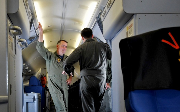 Two military personnel in olive/sage green flight suits are conversing inside what appears to be a military transport or VIP aircraft. The foreground features a blue seat with a black headrest cover embroidered with 'VIP' in red letters along with a military unit patch. The aircraft interior shows a narrow aisle with overhead compartments, blue carpeting, and equipment cases visible in the background.