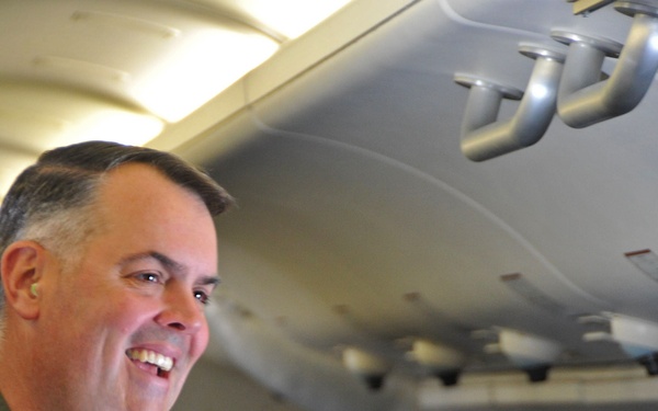 A smiling military officer wearing an olive green flight suit with patches is photographed inside what appears to be a military aircraft. The name tape on his patch reads 'Kevin Long' with the title 'Commodore.' A second person in a similar flight suit is partially visible on the right side of the frame.