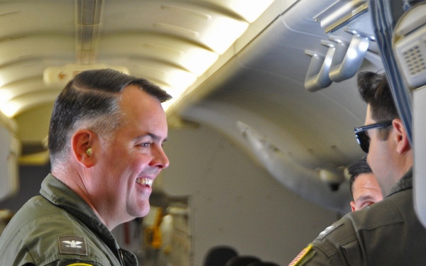 Military personnel in olive green flight suits are conversing inside what appears to be a military patrol aircraft. The individual on the left is smiling and wearing a patch identifying the P-8A Poseidon maritime patrol aircraft, along with a silver eagle rank insignia indicating a senior officer (Colonel or Navy Captain). The interior setting is consistent with the cabin of a P-8A Poseidon aircraft.