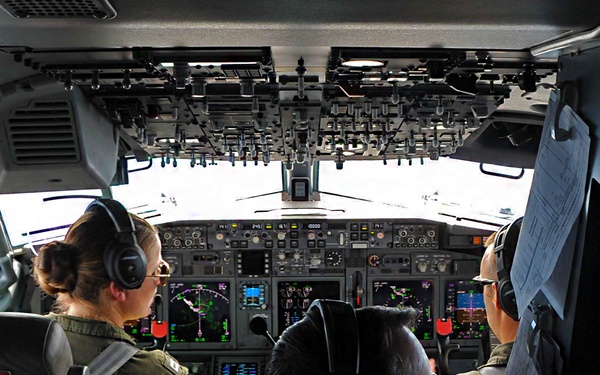 The image shows the interior of a military aircraft cockpit with three crew members wearing olive/green flight suits and aviation headsets. The cockpit features a modern glass cockpit display with multiple multifunction displays showing navigation and flight data. The aircraft appears to be on the ground or taxiing, visible through the forward windscreen.