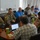 A mixed group of military personnel in camouflage uniforms and civilians in casual attire are gathered around a conference table in what appears to be an operations or briefing room. Multiple laptops are open and participants appear to be engaged in a collaborative working session or briefing. Green branded coffee cups are visible on the table, suggesting an extended working meeting.