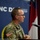 A U.S. Army soldier in MultiCam/OCP camouflage uniform is speaking at a podium with a microphone at an NC Department of Information Technology event. The soldier wears glasses and has an American flag patch on the left sleeve. The setting appears to be a formal briefing or press conference in North Carolina.