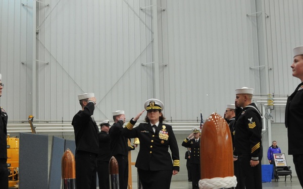 A U.S. Navy officer, appearing to be of senior rank based on gold sleeve stripes and multiple medals/ribbons, walks along a red carpet during a formal military ceremony inside a large industrial hangar. Personnel in Navy dress uniforms are lined up on both sides rendering salutes as the officer passes through. The ceremony appears to be a sideboy honors or change of command event, marked by decorative chrome bollards with rope and wooden finials flanking the red carpet.