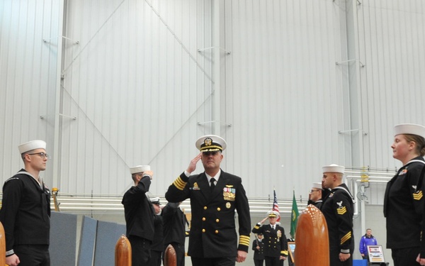 A U.S. Navy officer, wearing a dress uniform with gold insignia and service ribbons, walks along a red carpet during a formal military ceremony, likely a change of command or commissioning event. He is flanked by enlisted sailors in dress blue uniforms standing at attention or rendering salutes. The event is held inside a large industrial or hangar-type facility.