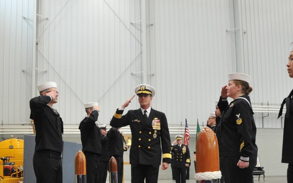 A U.S. Navy officer of senior rank walks along a red carpet through a sideboy honors ceremony inside a large industrial hangar facility. Enlisted sailors in dress blue uniforms and white Dixie cup covers are positioned on both sides of the red carpet, rendering salutes. Decorative chrome and wood bollards with rope lines flank the ceremonial pathway.