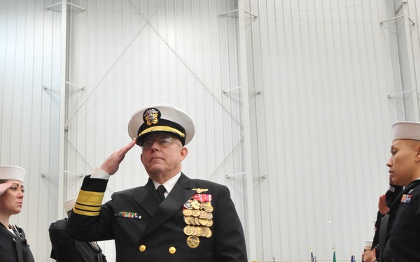 A formal U.S. Navy military ceremony is taking place inside a large hangar or indoor facility. A senior flag officer in dress blues with extensive medal and ribbon decorations is rendering a salute, flanked by two enlisted sailors also saluting. A color guard with flags is visible in the background on a red carpet.